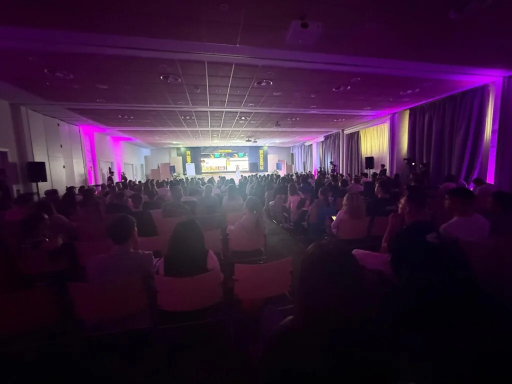 Sala de Billionaire 2.0 llena de asistentes con iluminación magenta y pantalla al fondo
